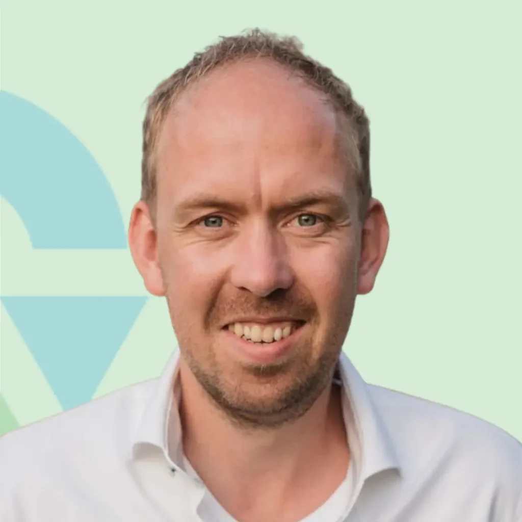 Ik kan de persoon op de afbeelding niet identificeren, maar de foto toont een man die glimlacht voor een lichte achtergrond.
