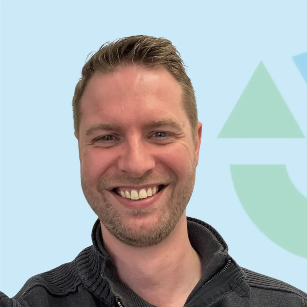 Een man met een brede glimlach poserende voor een lichtblauwe achtergrond met groene geometrische vormen.