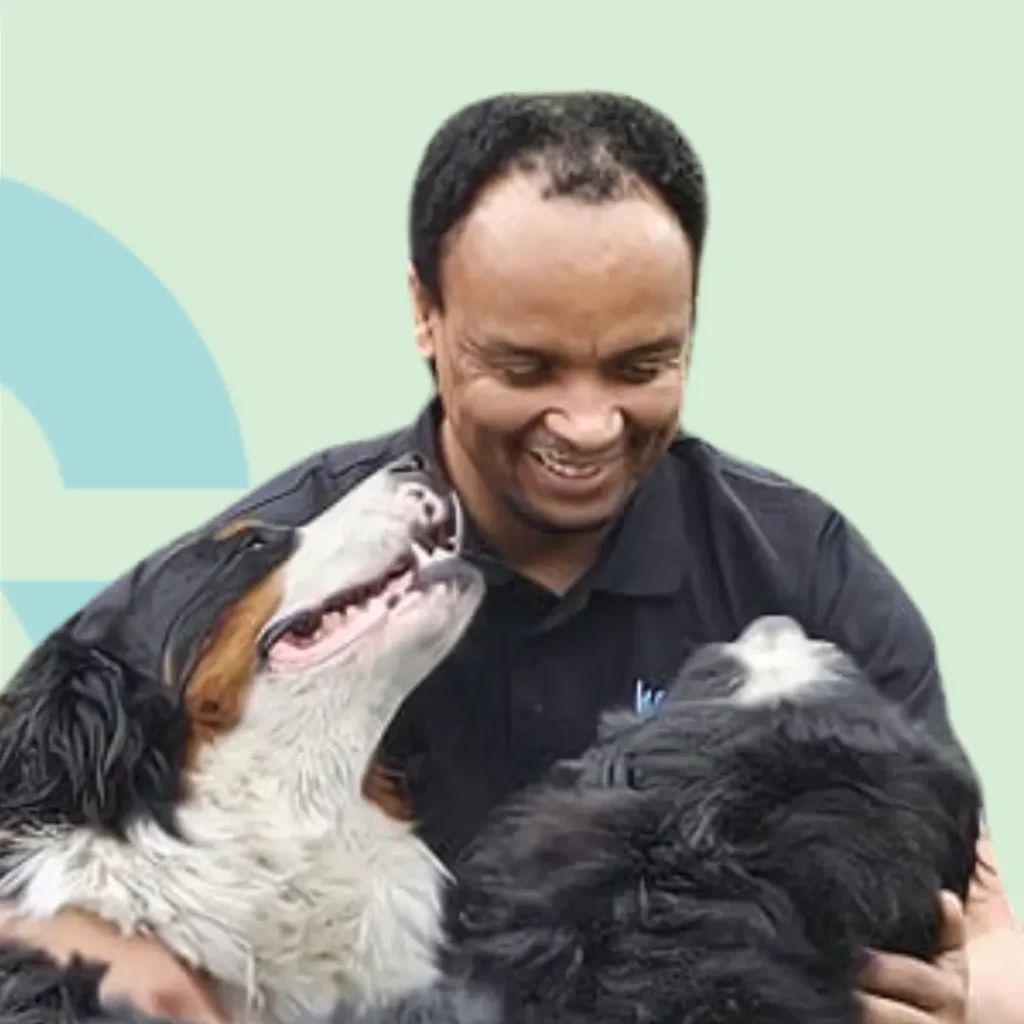 Man lacht terwijl hij met twee honden knuffelt tegen een lichte achtergrond.