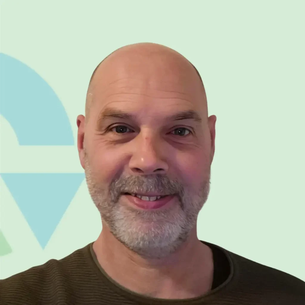 Ik weet niet wie de persoon is, maar de afbeelding toont een man met een baard en een glimlach, tegen een eenvoudige achtergrond.