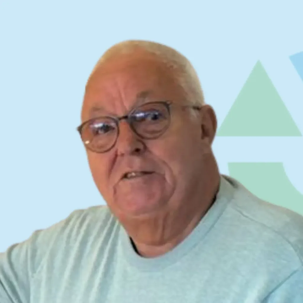 Een oudere man met een bril en grijs haar staat voor een lichtblauwe achtergrond.