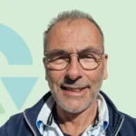 Een lachende man met een bril en kort haar, voor een lichtgroene achtergrond.