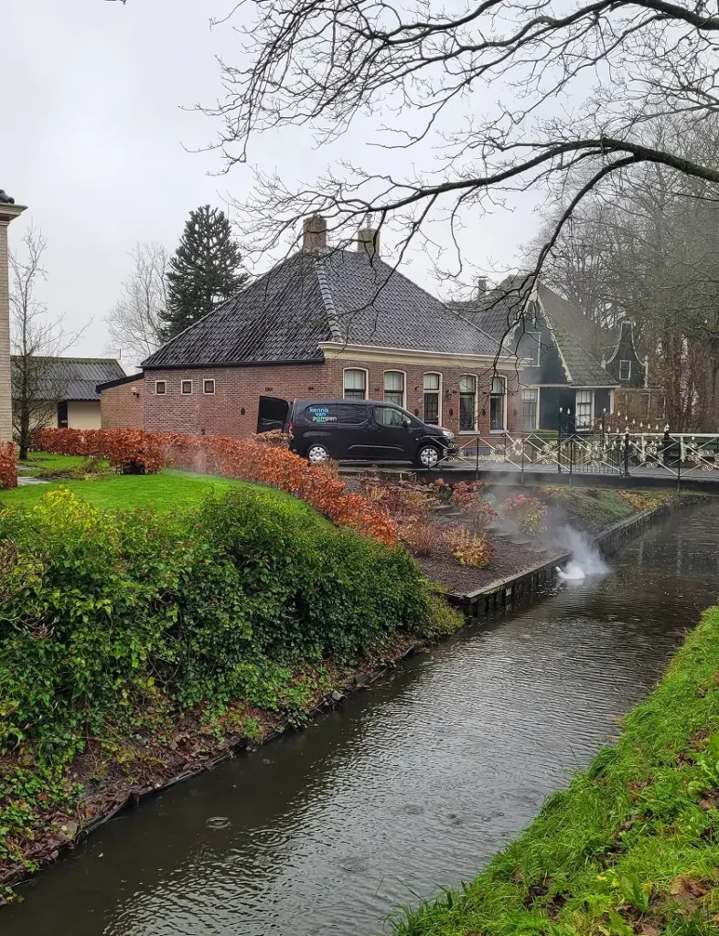 Een zwart bedrijfsbusje staat geparkeerd naast een traditionele woning terwijl rook uit een naastgelegen sloot komt, mogelijk in verband met het opsporen van hemelwateraansluitingen op het drukriool.