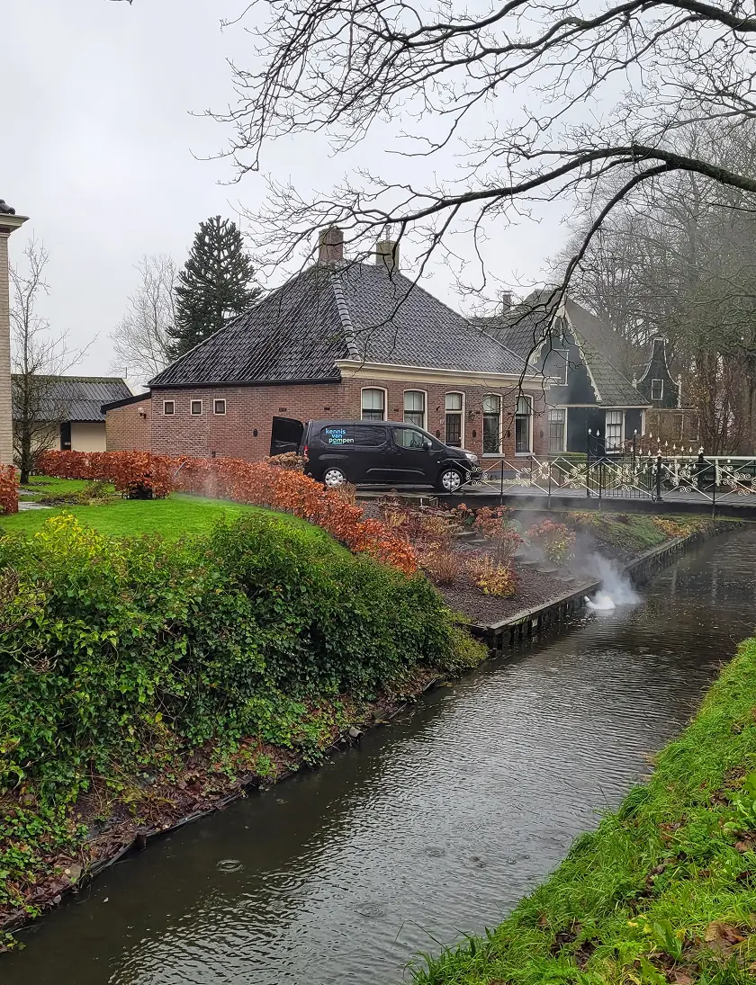 Een zwart bedrijfsbusje staat geparkeerd naast een traditionele woning terwijl rook uit een naastgelegen sloot komt, mogelijk in verband met het opsporen van hemelwateraansluitingen op het drukriool.