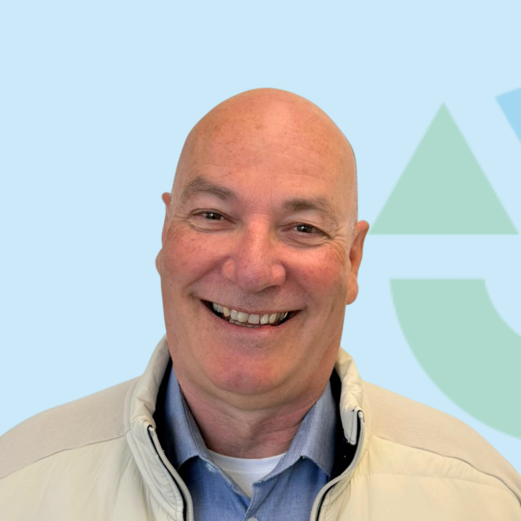 Een lachende man met een kaal hoofd en een lichte jas, voor een blauwgroene achtergrond met geometrisch patroon.