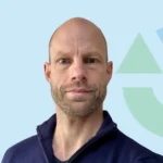 Portret van een man met een lichte baard en een navy trui voor een neutrale achtergrond.