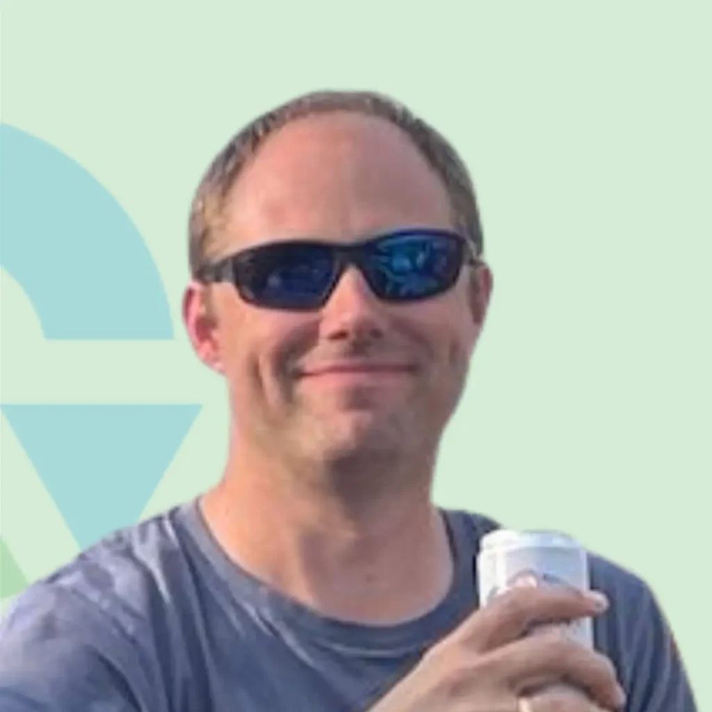 Iemand met een zonnebril en glimlach houdt een drankje vast tegen een lichte achtergrond.