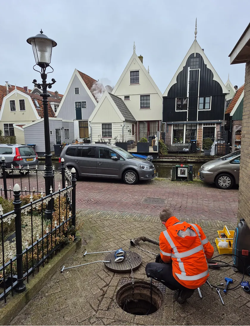 Een technicus voert een nul-inspectie uit bij een open riooldeksel in een pittoreske woonwijk met traditionele Nederlandse huizen.