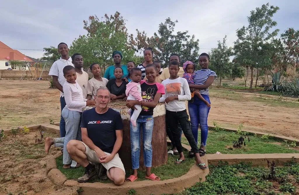 Groepsfoto van een glimlachende man en een groep kinderen in een buitenomgeving van Tanzania met bomen en gras.