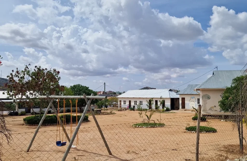 Speeltuin in Tanzania met een schommel voor een omheind gebouw onder een bewolkte hemel in een droge omgeving.