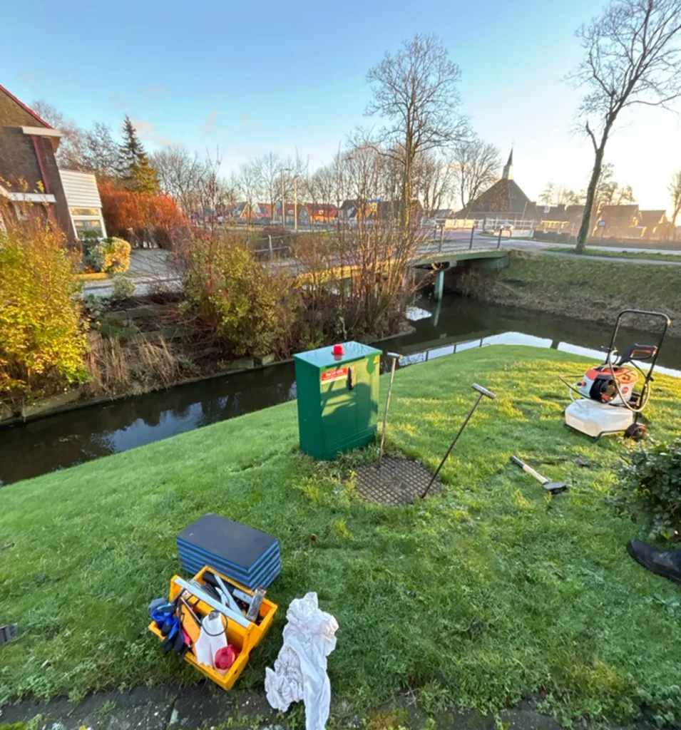 Grasveld langs een sloot met gereedschap en een grasmaaier bij een elektriciteitskast in een landelijke omgeving.