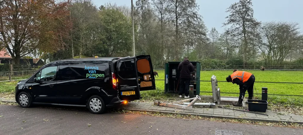 Monteurs van "Kennis van pompen" werken aan een installatie langs een groen veld, met hun bedrijfsbus geparkeerd op de stoep.