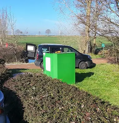 Een groene elektriciteitskast op het gras naast een bedrijfsbusje met open achterdeur in een landelijke omgeving.