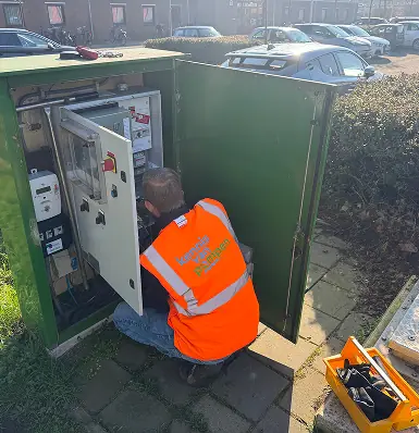 Een technicus in een oranje hesje voert onderhoud uit aan een open verdeelkast op straat.