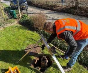 Een man in een oranje veiligheidshes inspecteert een put op een grasveld.