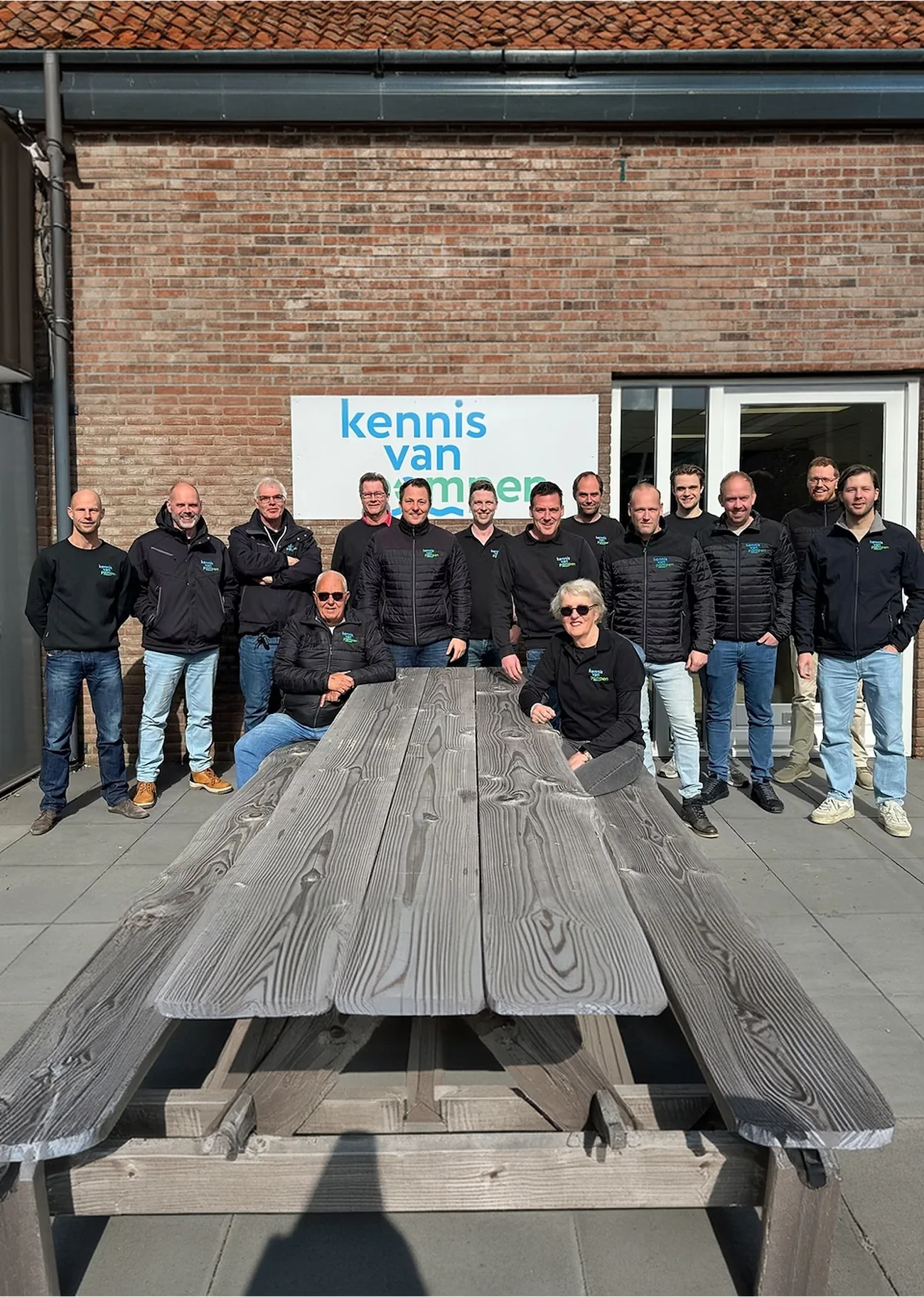 Teamfoto van medewerkers bij een bord met "kennis van pompen" op een zonnig terras.