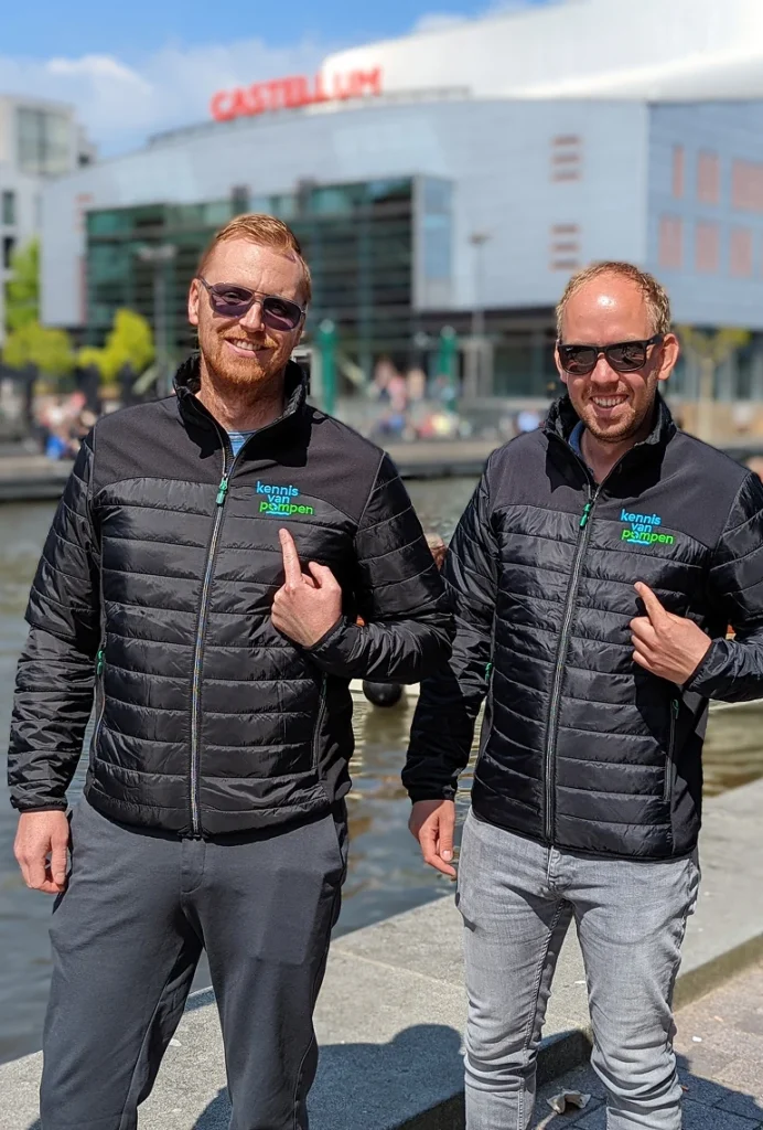 Twee mannen in jassen met het logo 'kennis van pompen', poseren voor een modern gebouw naast water.